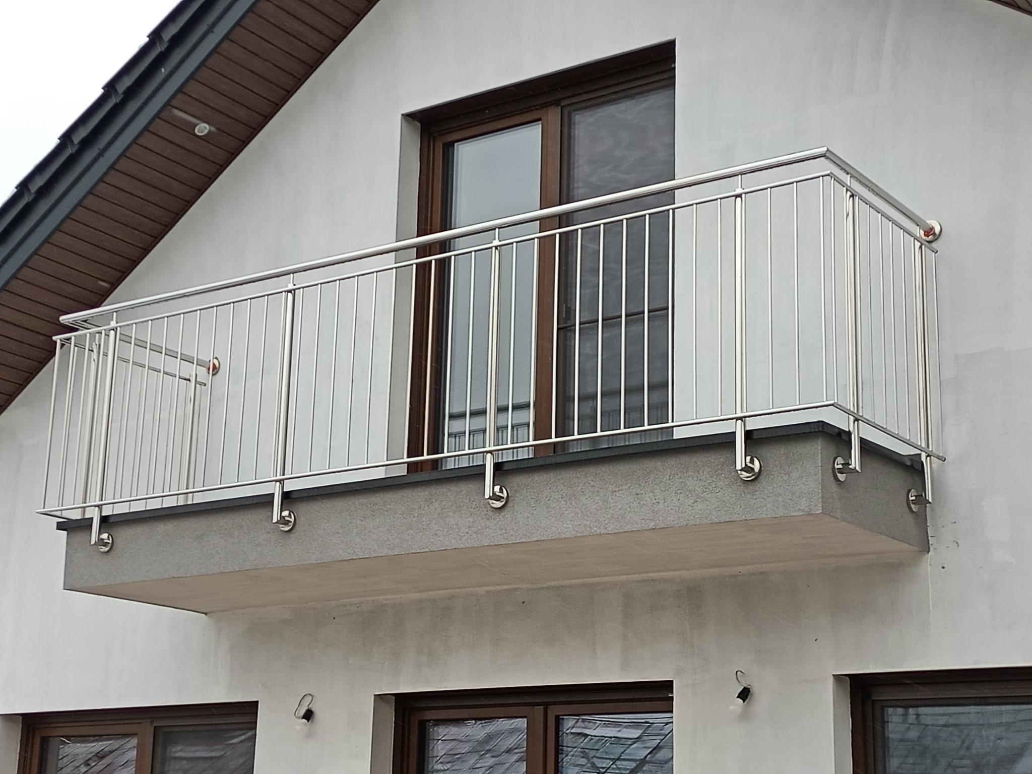 Balkon z nierdzewną balustradą, widok z dołu. Metalowe słupki i poręcze, mocowane do betonowej płyty balkonu. Okno balkonowe w tle. Oświetlenie zewnętrzne zamontowane pod balkonem.