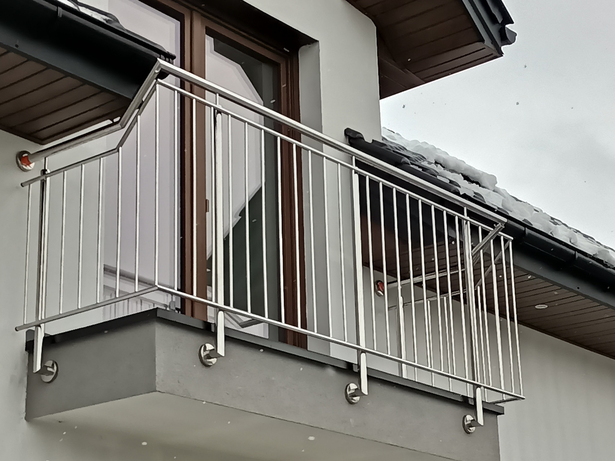 Nowoczesna, srebrna balustrada balkonowa ze stali nierdzewnej, mocowana na zewnątrz budynku. Widoczne detale mocowania do ściany i odbicie w szybie okiennej.