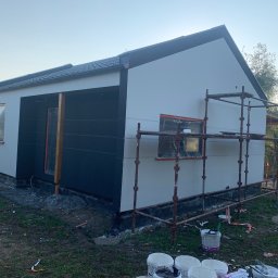 PAT-BUD - Elewacja domu w trakcie malowania: część ściany biała, część grafitowa. Okna zabezpieczone folią, rusztowanie, puszki z farbą. Widok z zewnątrz, w tle zieleń.