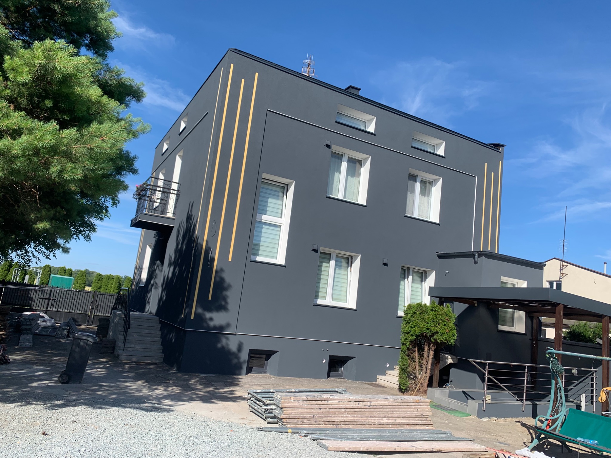 Elewacja domu po malowaniu na grafitowo, z żółtymi pionowymi pasami dekoracyjnymi. Widoczne okna z białymi ramami i niebieskie niebo w tle.