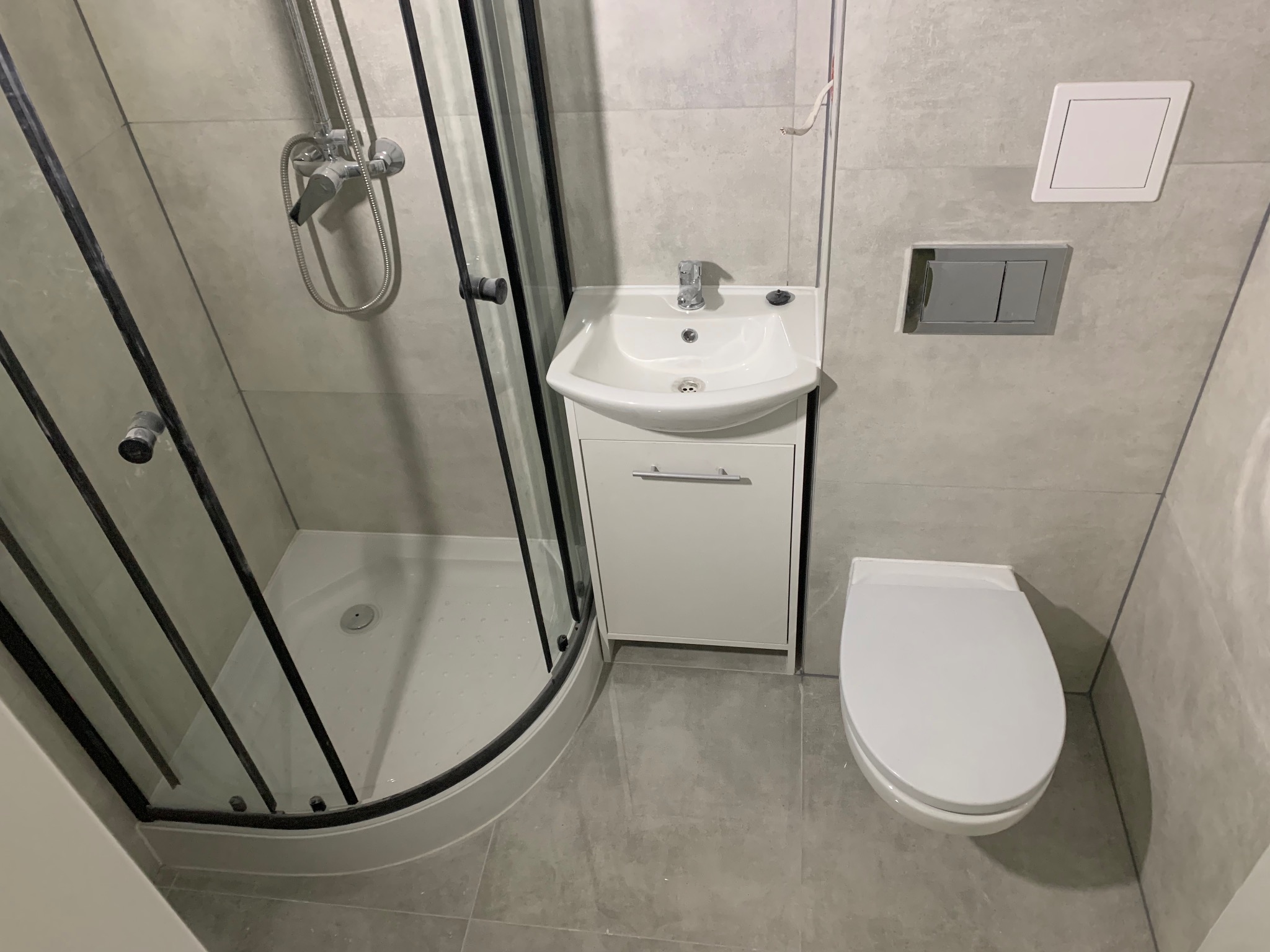 Nowoczesna łazienka z kabiną prysznicową narożną, umywalką z szafką i podwieszaną toaletą. Szare płytki tworzą minimalistyczny design.