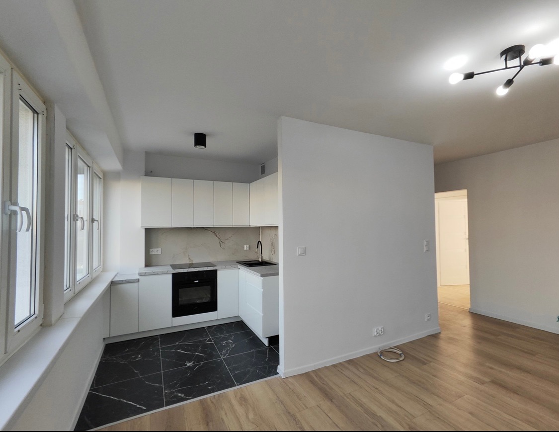 Nowoczesne wnętrze: biała kuchnia z czarnym blatem i podłogą, drewniana podłoga w salonie, minimalistyczne oświetlenie, jasne ściany i okna.