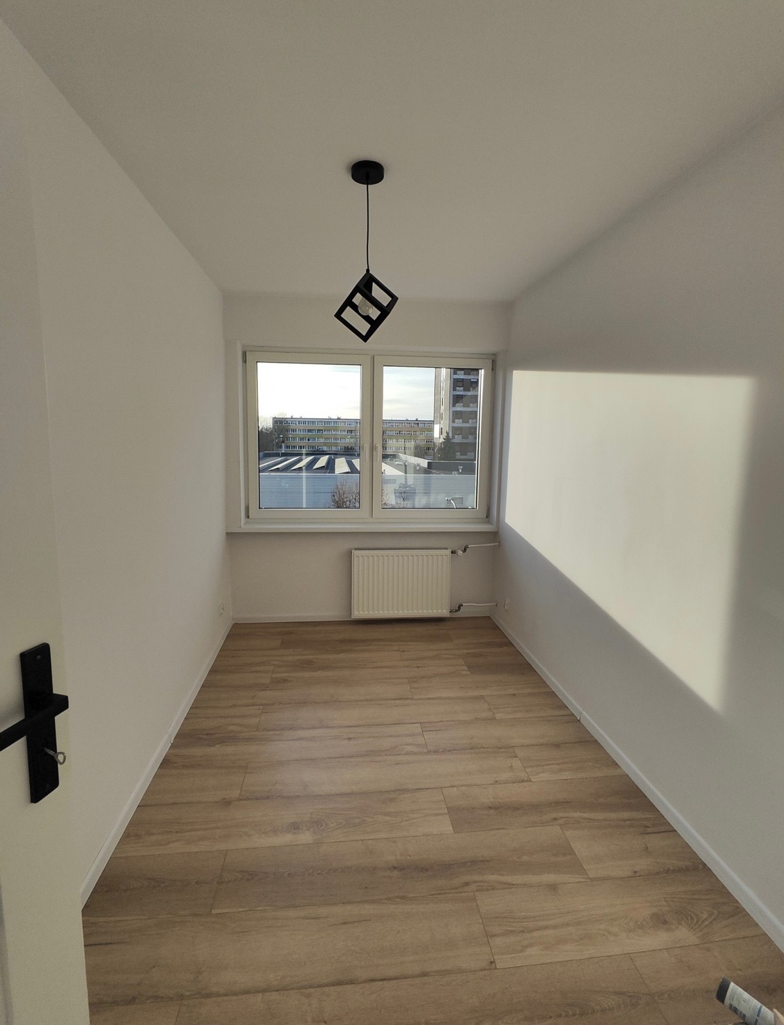 Jasne, świeżo wykończone wnętrze pokoju z drewnianą podłogą, białymi ścianami i nowoczesną lampą kostką. Widok z okna na budynki.