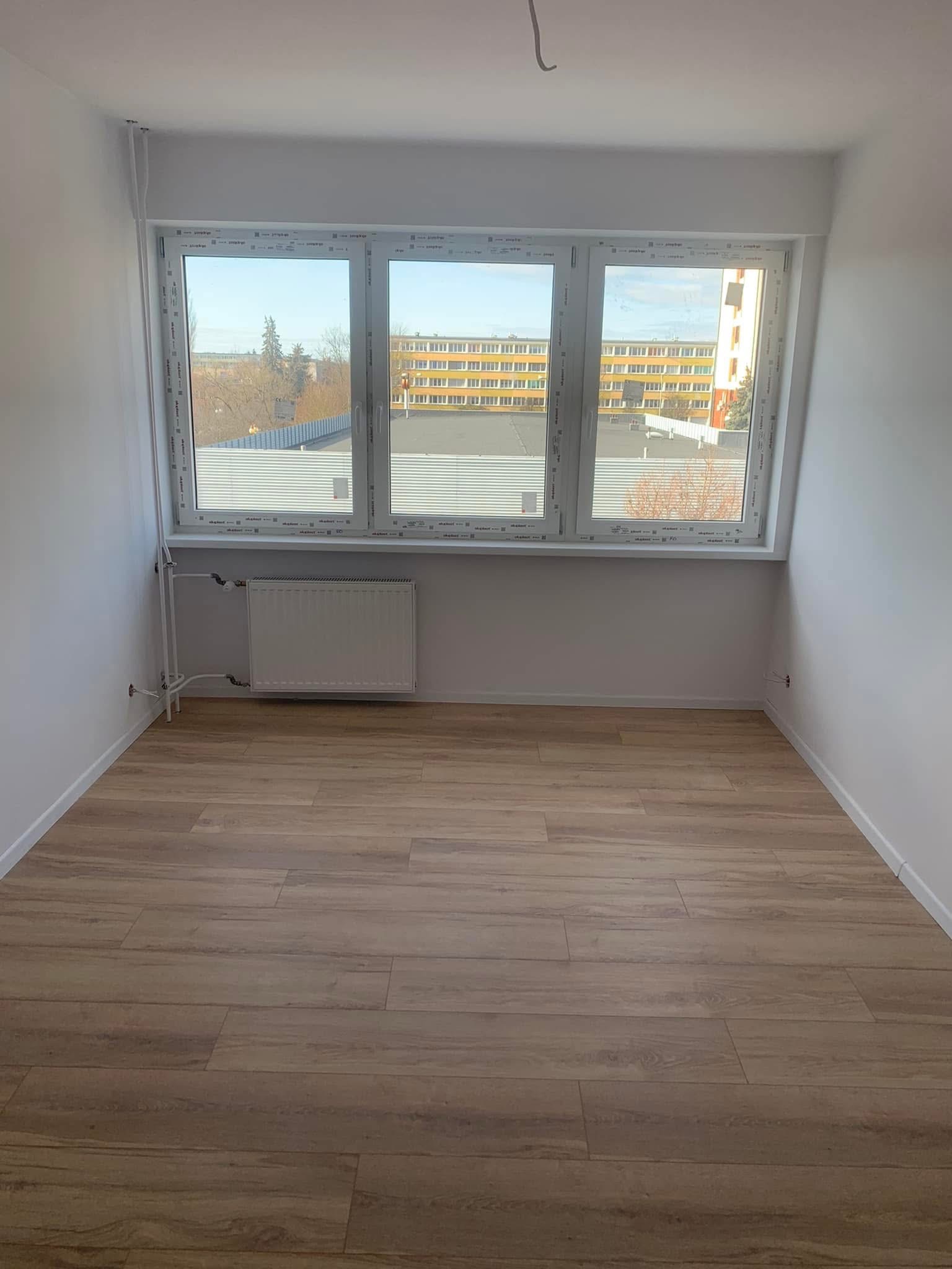 Jasny pokój po remoncie: świeżo pomalowane ściany, ułożona podłoga z paneli imitujących drewno, białe okno z widokiem na budynek. Grzejnik pod oknem.