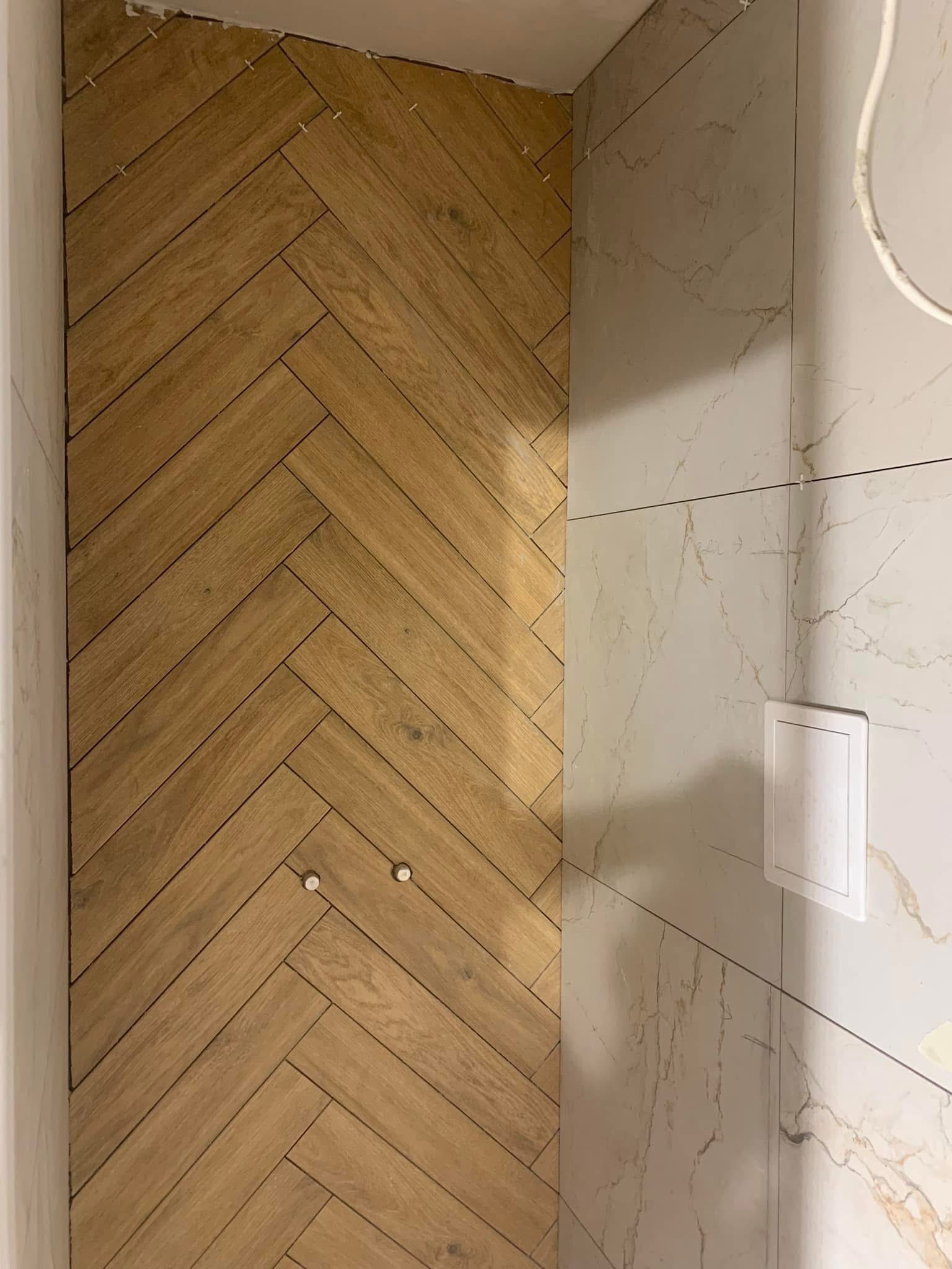 Wykończona ściana łazienki z drewnopodobnymi płytkami ułożonymi w jodełkę i marmurowymi płytkami. Widoczne krzyżyki dystansowe.