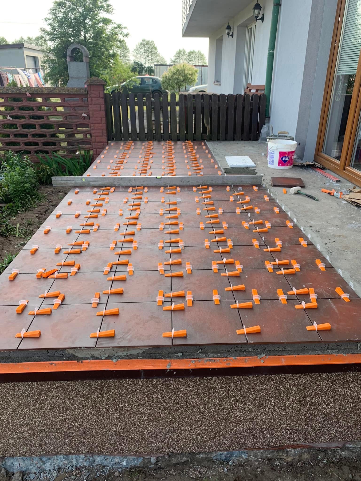 Układanie brązowych płytek na tarasie z użyciem pomarańczowych krzyżyków dystansowych. Widoczny biały pojemnik z klejem Atlas i poziomica.