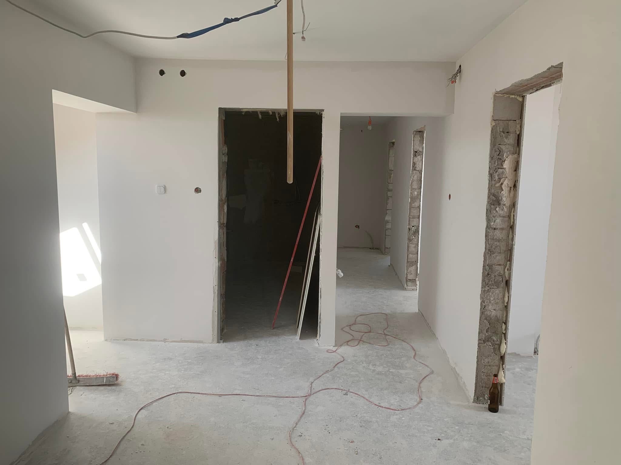 Wnętrze w trakcie remontu: świeżo pomalowane ściany na biało, surowy beton na podłodze, widoczne otwory drzwiowe, kable na suficie, butelka przy ścianie.