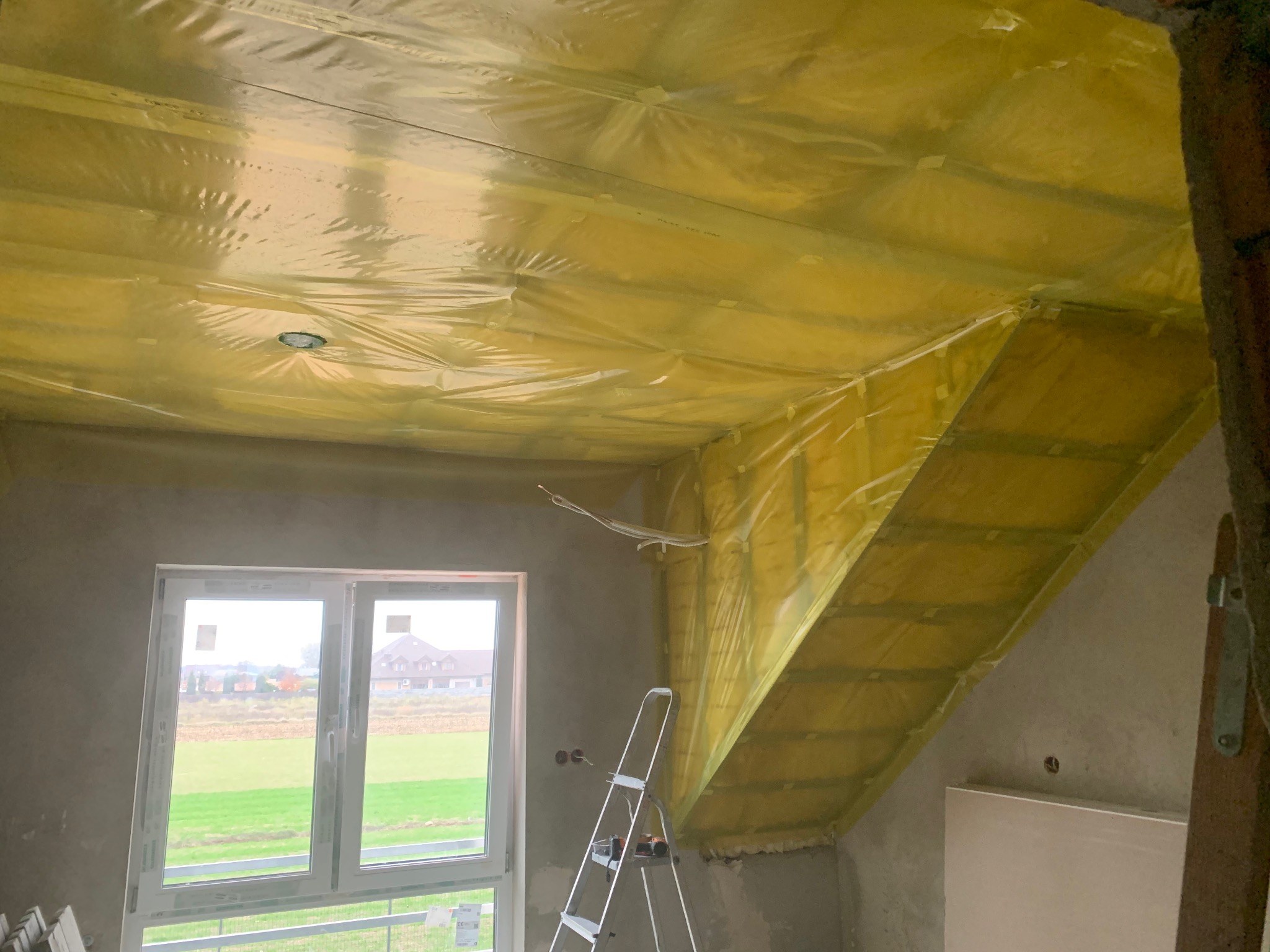 Ocieplenie poddasza folią paroizolacyjną, widoczna drabina, okno i surowy stan ścian. Prace przygotowawcze przed wykończeniem wnętrza na poddaszu.