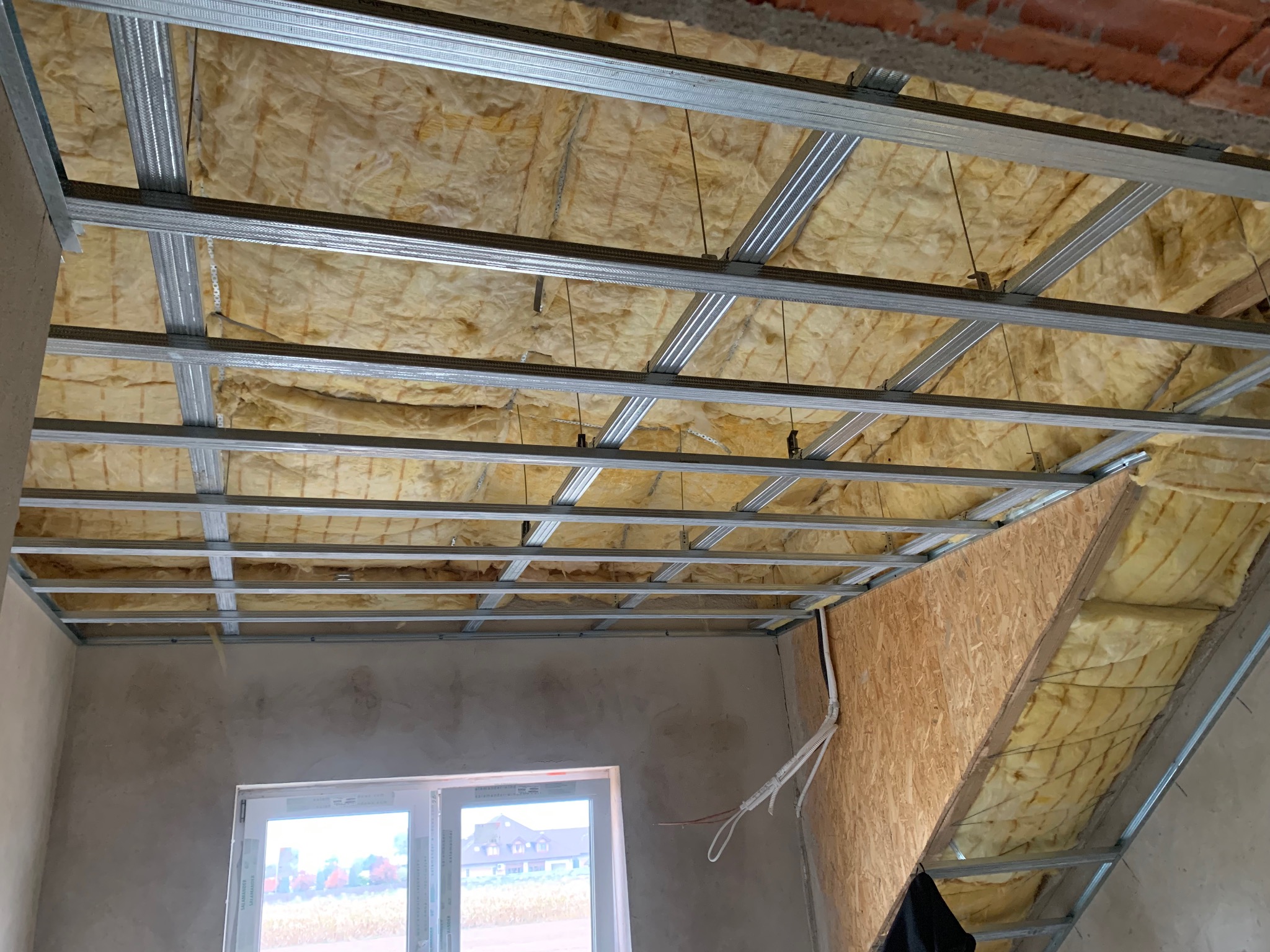 Sufit podwieszany z izolacją z wełny mineralnej i metalową konstrukcją, widok z dołu, w tle okno i fragment ściany. Prace wykończeniowe na poddaszu.