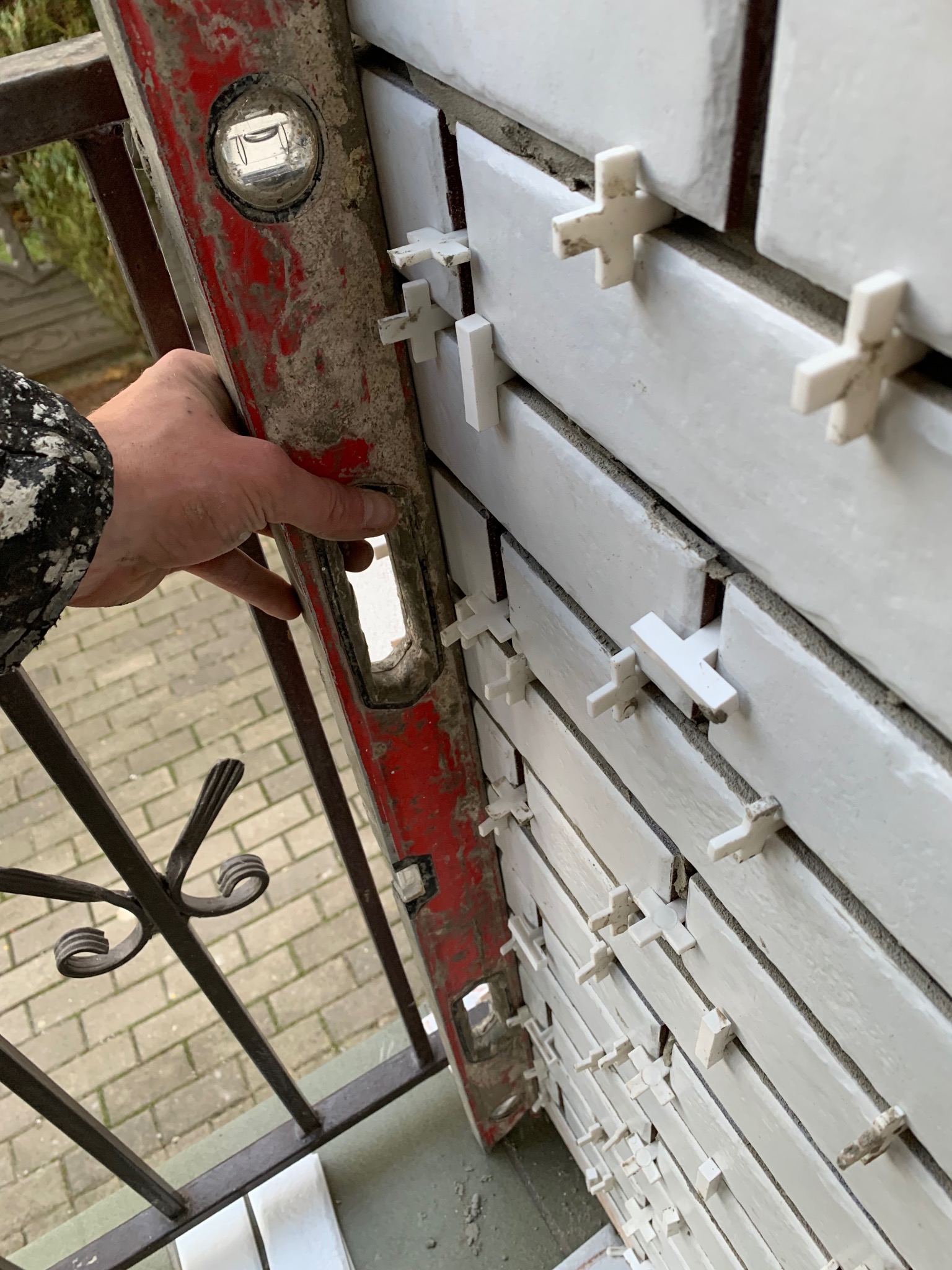 Układanie białych płytek na balkonie z użyciem krzyżyków dystansowych, sprawdzane poziomicą. Widoczne barierki balkonu i fragment podłogi.