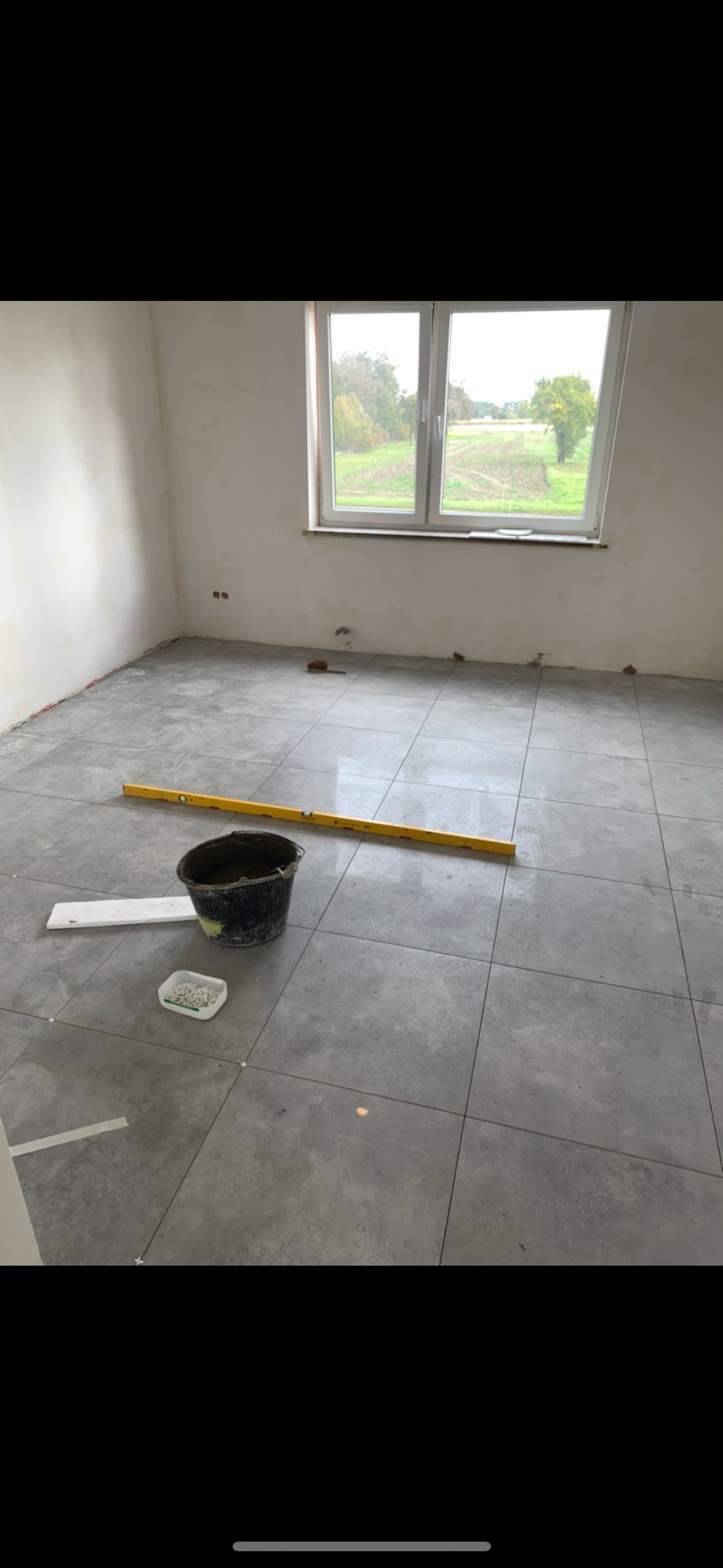 Szare płytki podłogowe w pokoju z oknem. Widoczny kubeł, poziomica i krzyżyki dystansowe. Prace wykończeniowe w trakcie układania płytek.