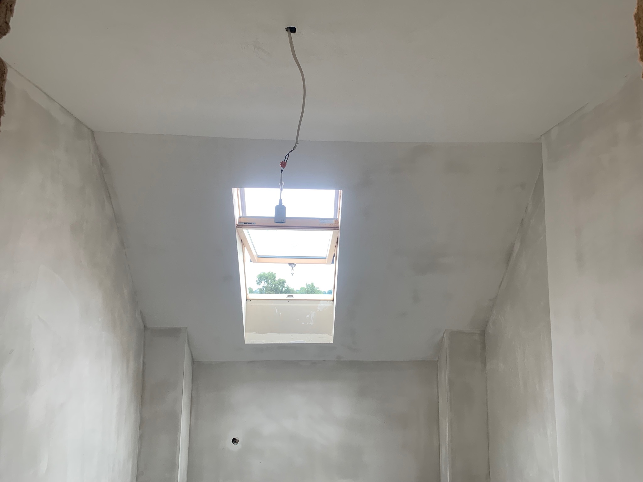Pokój ze ścianami po szpachlowaniu, widoczne okno dachowe i wystający kabel z sufitu. Przygotowanie do dalszych prac wykończeniowych.