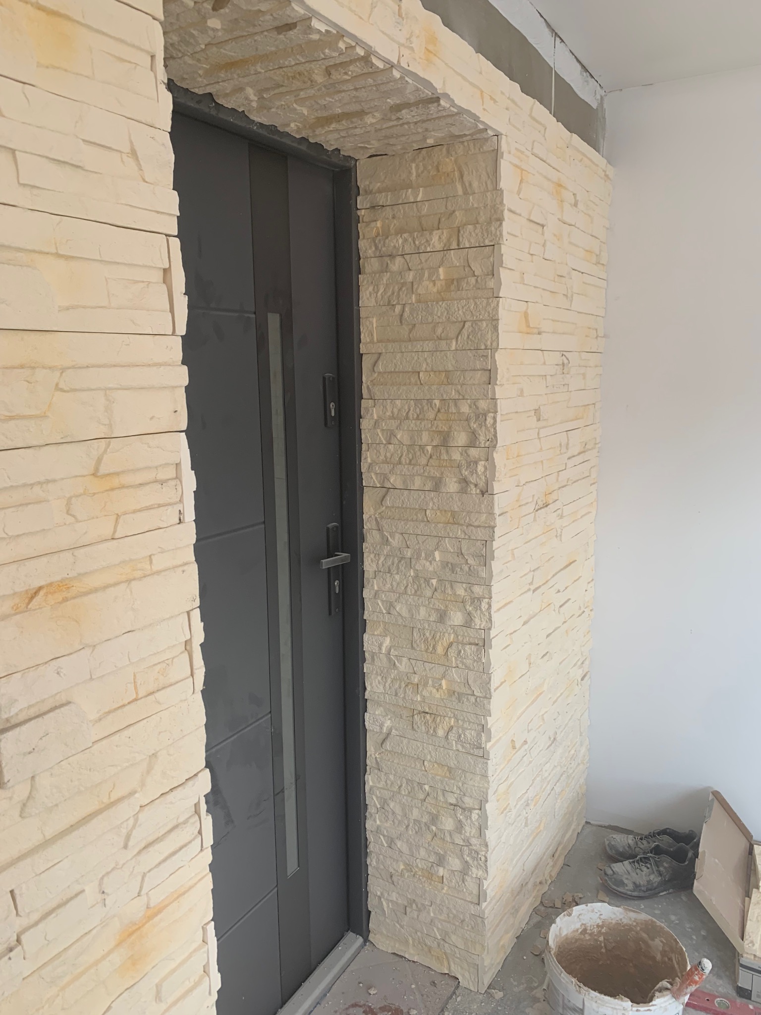 Wykończone obramowanie drzwi wejściowych z dekoracyjnej, beżowej cegły. W tle widoczne buty robocze i wiadro z zaprawą.
