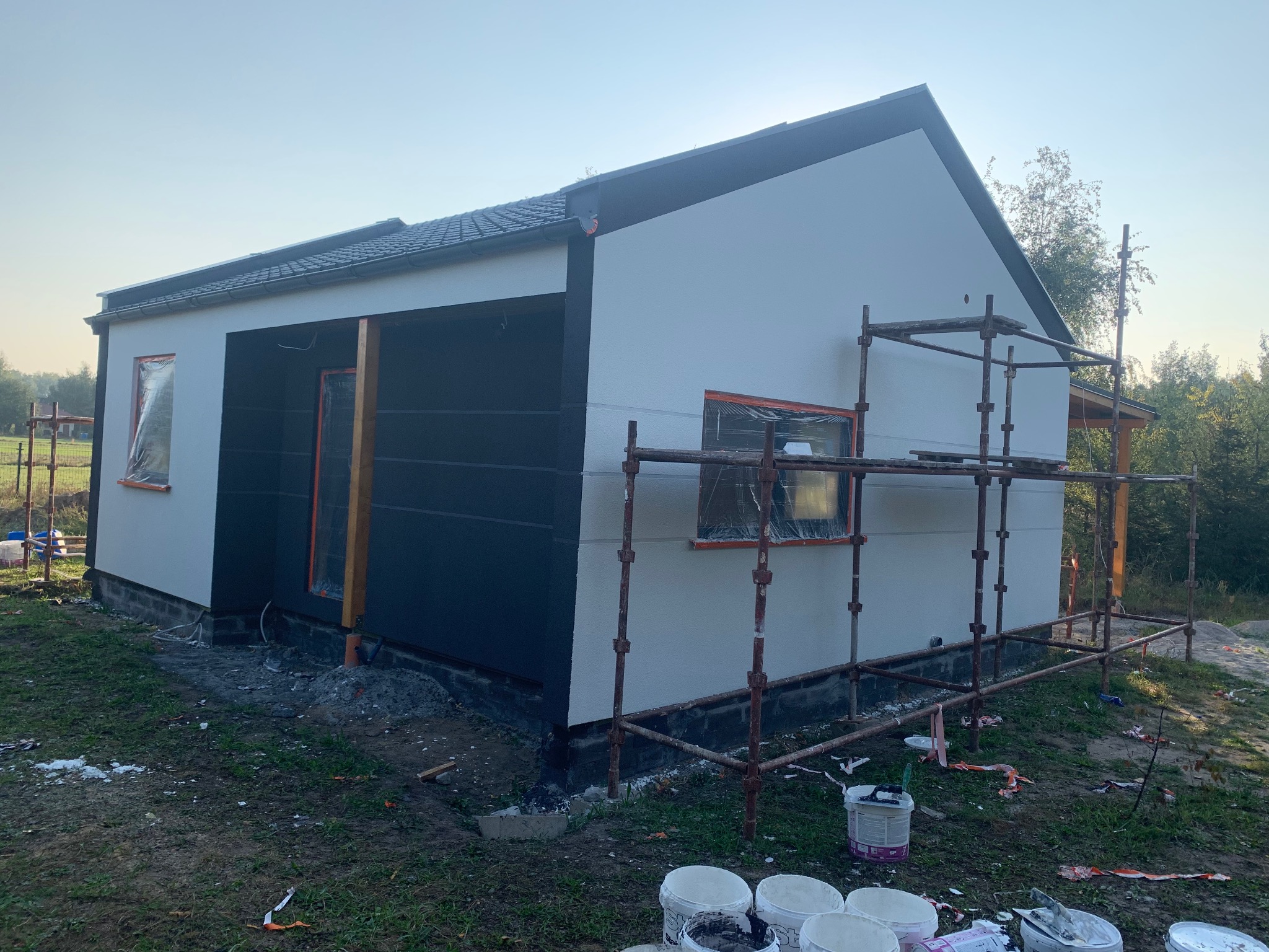 Elewacja domu w trakcie malowania: część ściany biała, część grafitowa. Okna zabezpieczone folią, rusztowanie, puszki z farbą. Widok z zewnątrz, w tle zieleń.