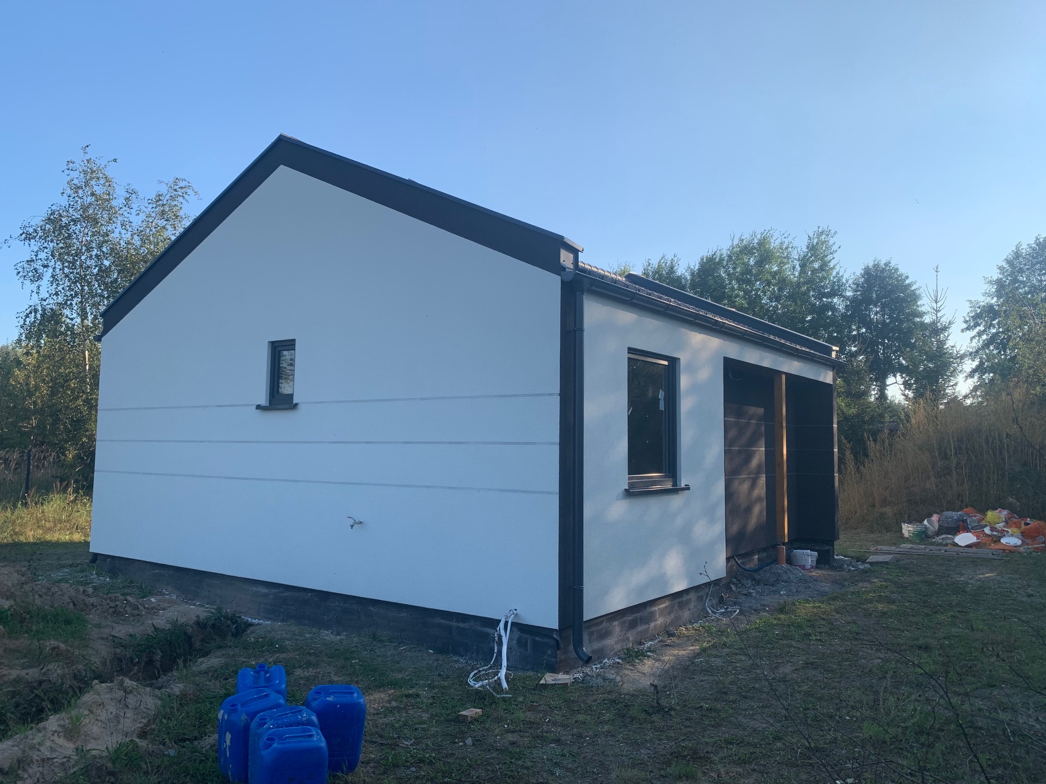 Biały budynek z szarymi detalami wykończeniowymi, okna w szarych ramach, widoczne niebieskie pojemniki i śmieci na trawie. Budynek w trakcie wykończenia.