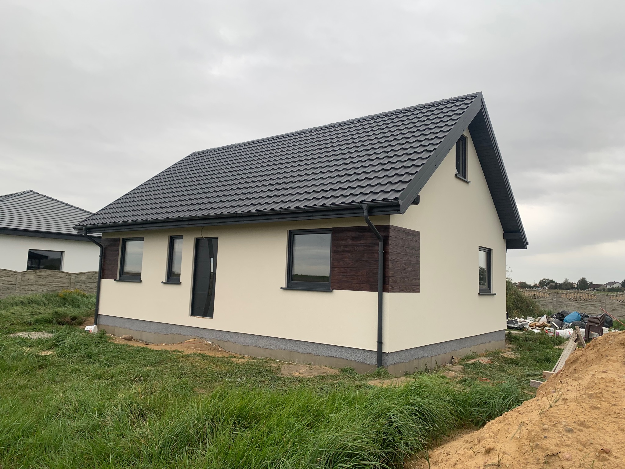 Nowo wybudowany dom jednorodzinny z beżową elewacją, grafitowym dachem i brązowymi panelami dekoracyjnymi. Widok z zewnątrz w pochmurny dzień.