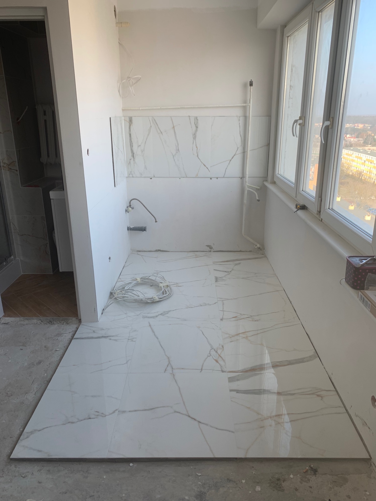 Biała łazienka z marmurowymi płytkami podłogowymi i ściennymi w trakcie wykańczania. Widoczne okno z widokiem na miasto i instalacja wodna.