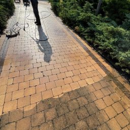 Myjto.pl - Czyszczenie kostki brukowej z użyciem myjki ciśnieniowej. Widoczna różnica między umytą a zabrudzoną częścią. Praca w toku, osoba w czarnym ubraniu obsługuje urządzenie.