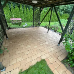 Zadaszona altana z nowo ułożoną, beżową kostką brukową. W tle zielony trawnik, rząd tuj i drewniana ławka. Widok z góry, równomierne oświetlenie.