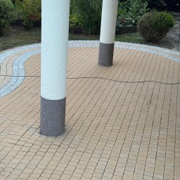 Myjto.pl - Beżowa kostka brukowa ułożona w okrąg z dekoracyjną obwódką, otaczająca dwie białe kolumny z szarymi podstawami. Tło stanowią krzewy i drzewa.