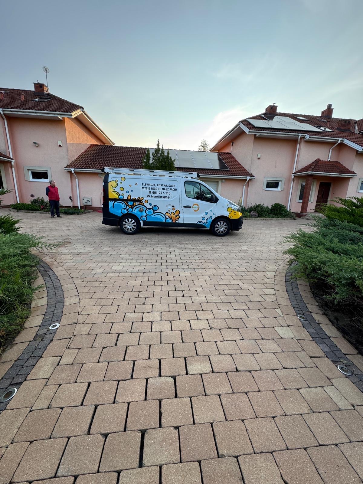 Brukowa aleja przed domami, firmowy van z ofertą czyszczenia elewacji i kostki, osoba w oddali. Kostka brukowa w odcieniach beżu, oświetlenie wzdłuż ścieżki.