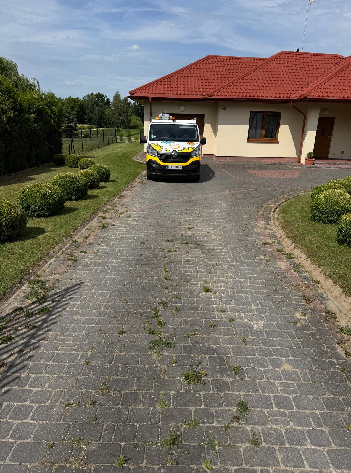 Kostka brukowa z chwastami przed domem jednorodzinnym. Na podjeździe stoi firmowy bus z żółtym pasem. Widok na jasną elewację domu z czerwoną dachówką.