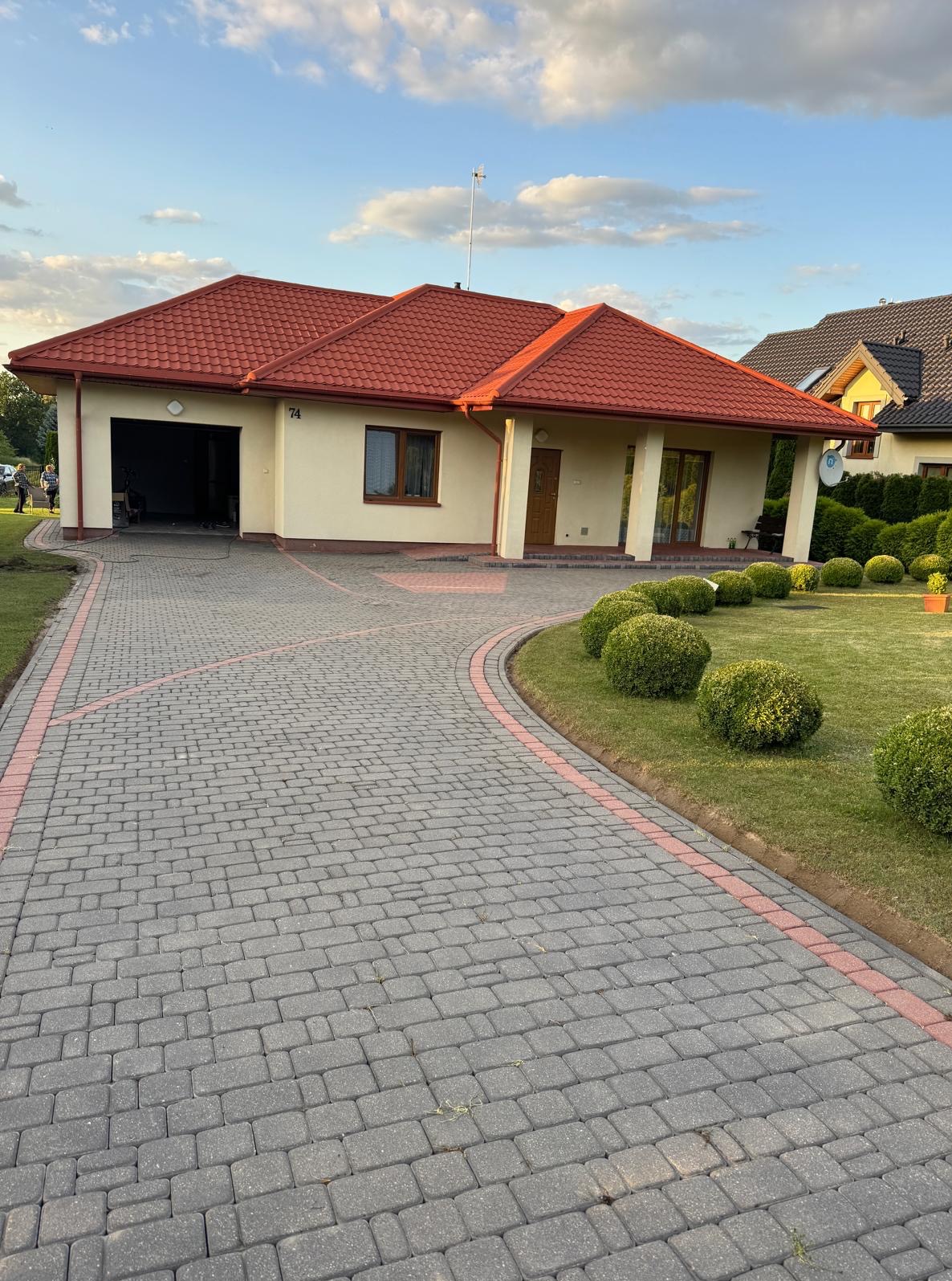 Dom z beżową elewacją i czerwoną dachówką, z widocznym podjazdem z kostki brukowej w odcieniach szarości z czerwoną obwódką. Zadbany trawnik z krzewami.