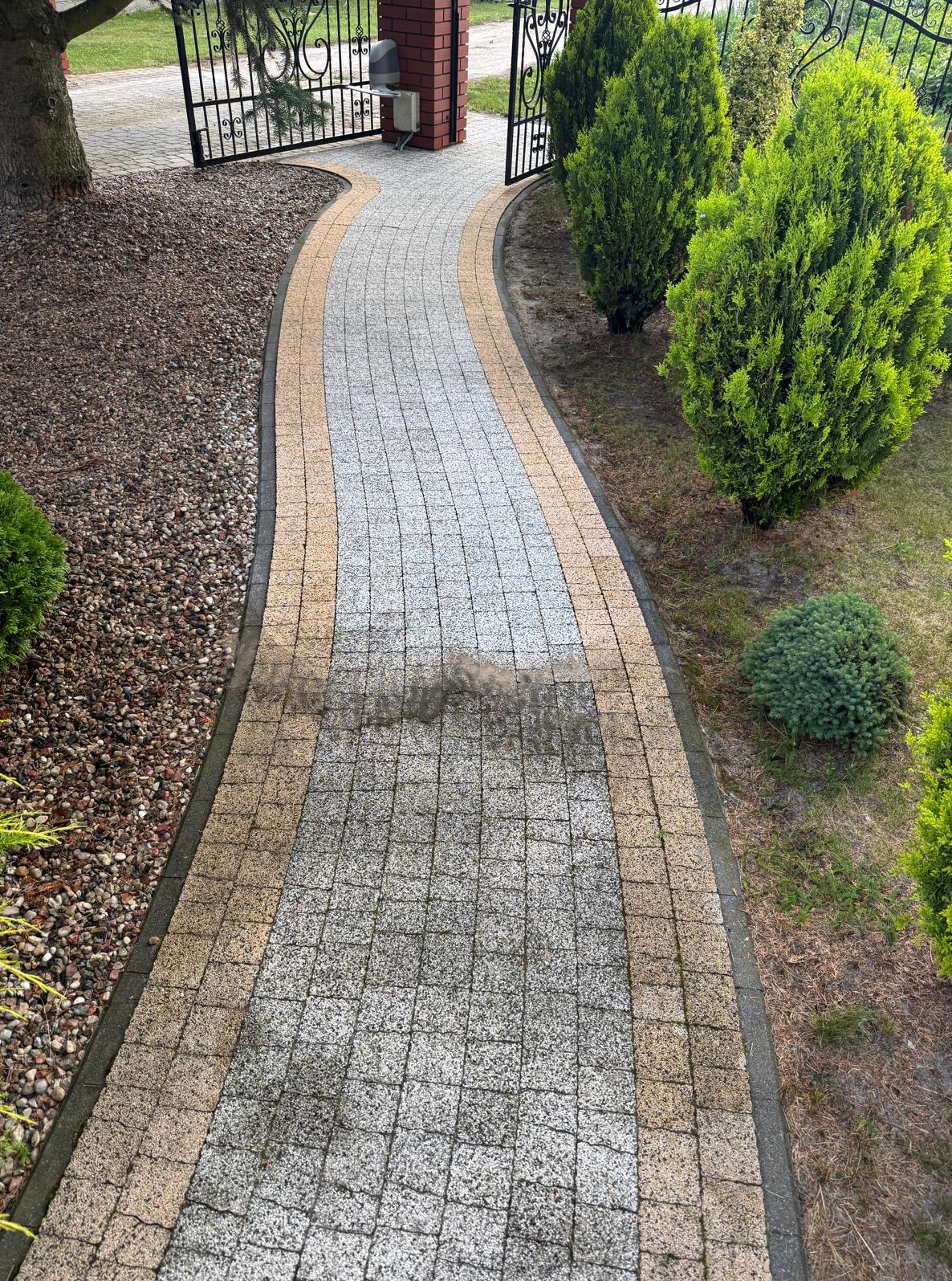 Fragment czyszczonej kostki brukowej w Lublinie, widoczne różnice w kolorze przed i po zabiegu. Ścieżka prowadzi w stronę bramy wjazdowej. Roślinność po bokach.