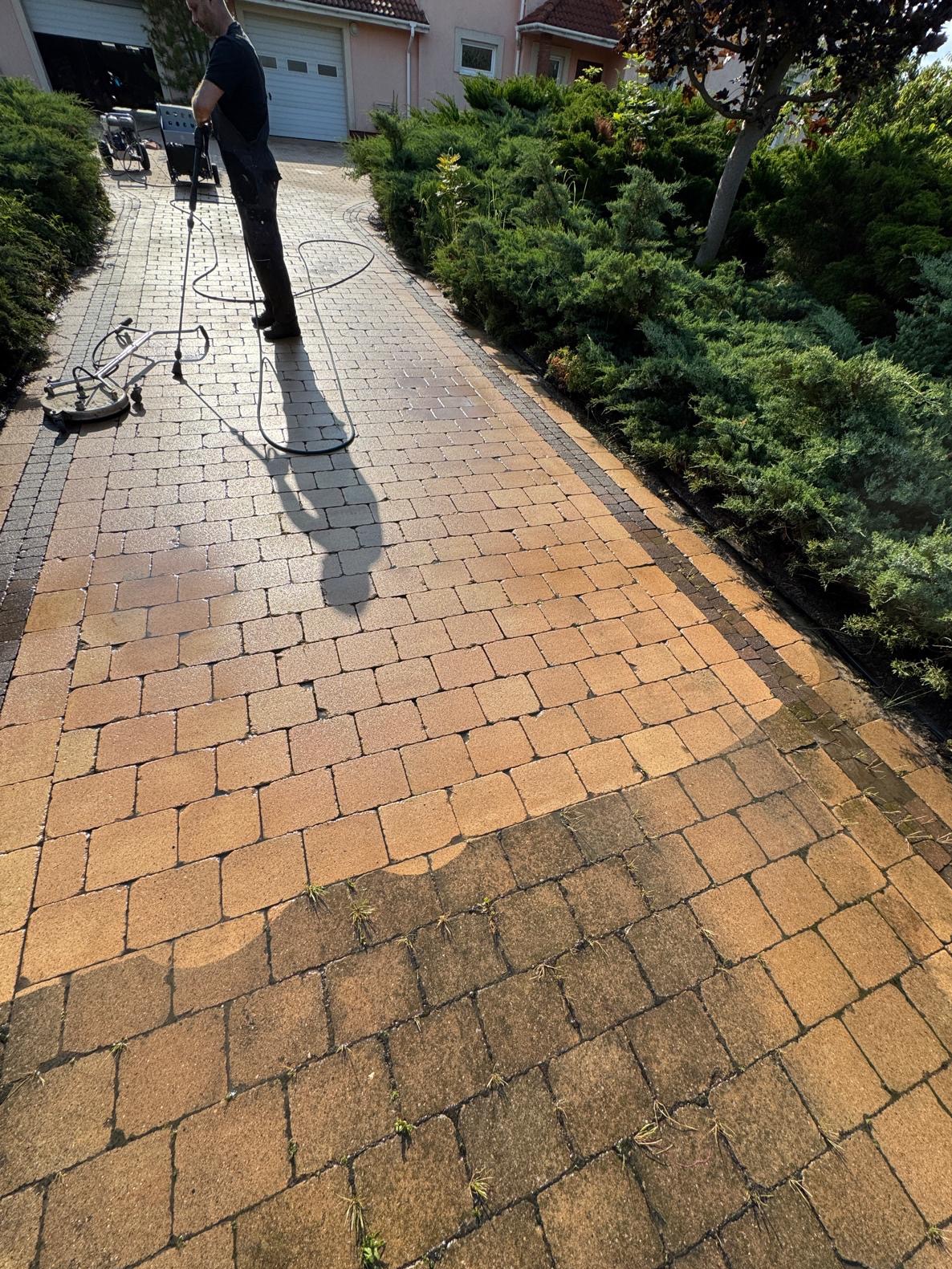 Czyszczenie kostki brukowej z użyciem myjki ciśnieniowej. Widoczna różnica między umytą a zabrudzoną częścią. Praca w toku, osoba w czarnym ubraniu obsługuje urządzenie.