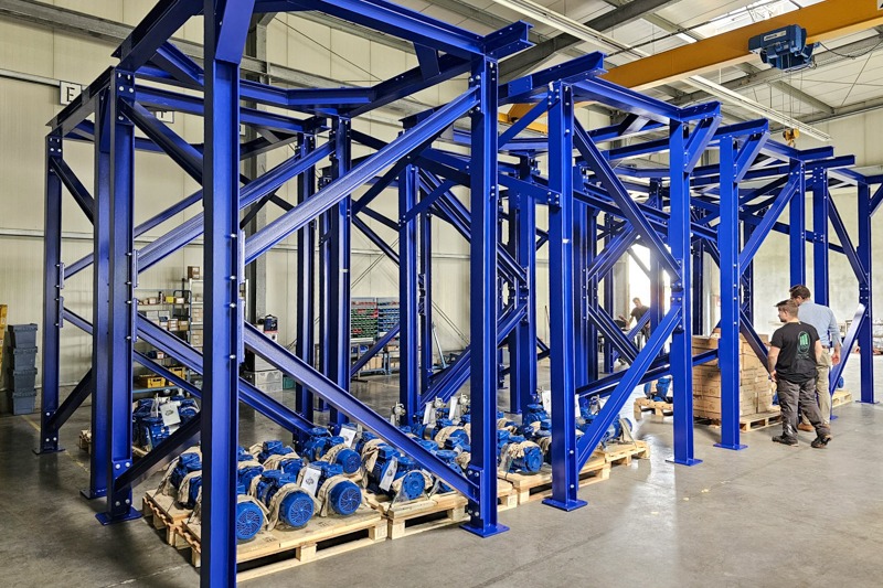 Niebieska, stalowa konstrukcja hali magazynowej z elementami na paletach. Widoczni pracownicy, suwnica i oświetlenie hali. Konstrukcja po malowaniu proszkowym.