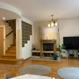 Jasne wnętrze domu z drewnianymi schodami, kominkiem i telewizorem. Parkiet na podłodze, okrągłe dywany. Widok na schody i przeszkloną ścianę.