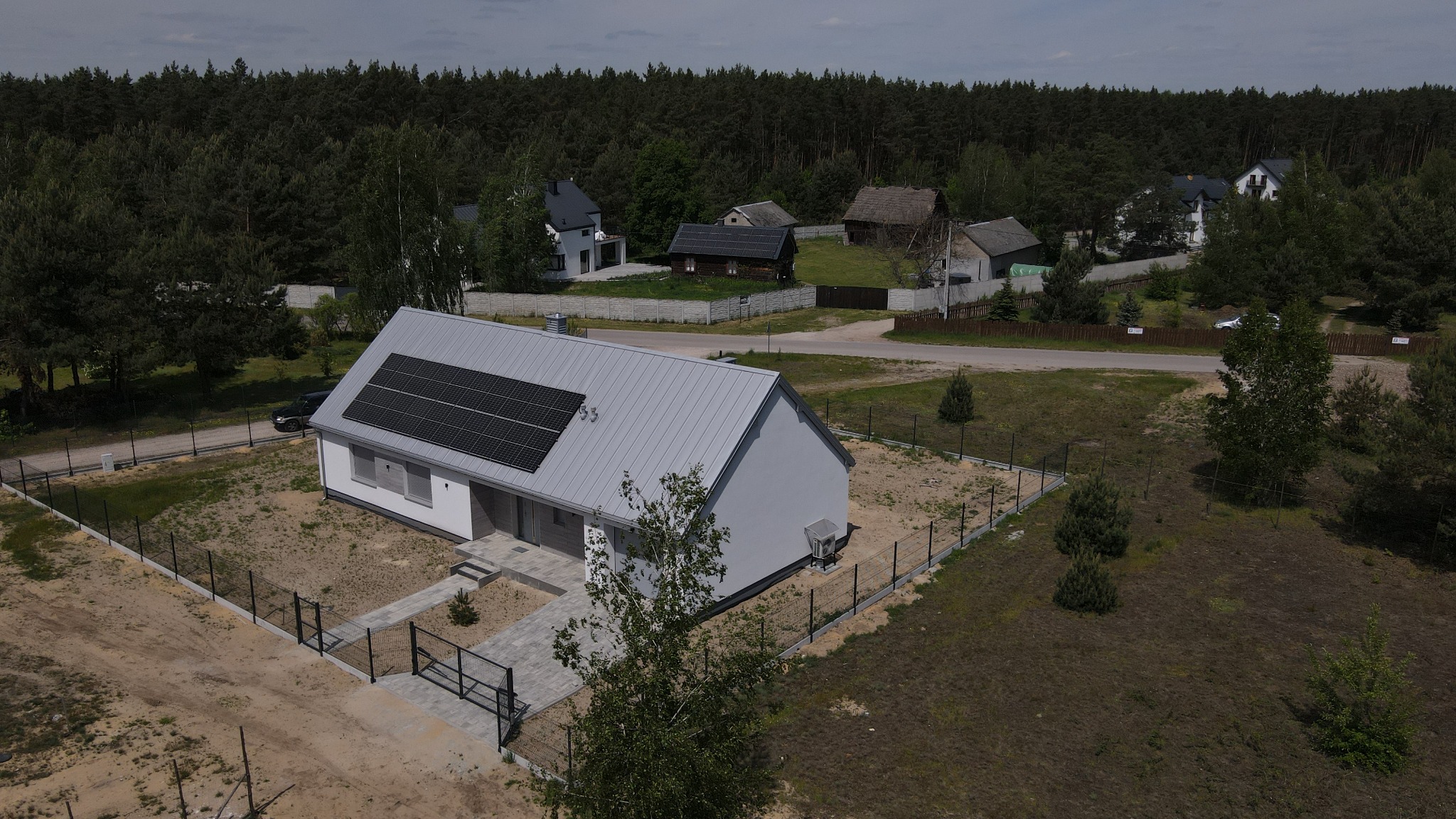 Nowoczesny, parterowy dom z szarą blachodachówką i panelami solarnymi na dachu, otoczony surowym terenem, widziany z lotu ptaka na tle lasu.