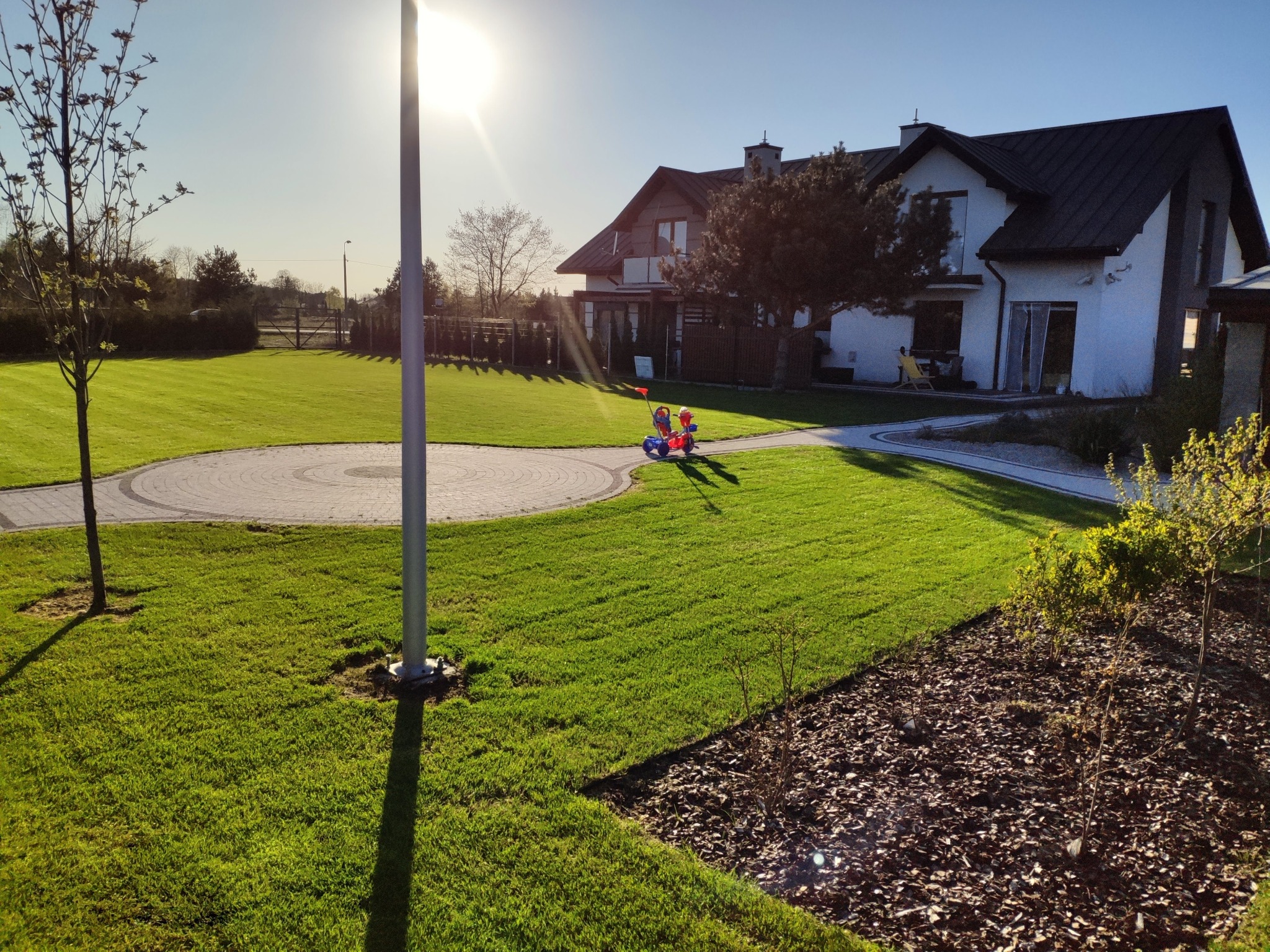 Nowoczesny dom z zadbanym trawnikiem, ścieżką z kostki i placem zabaw. Słoneczny dzień, błękitne niebo. W tle widoczny las i ogrodzenie.