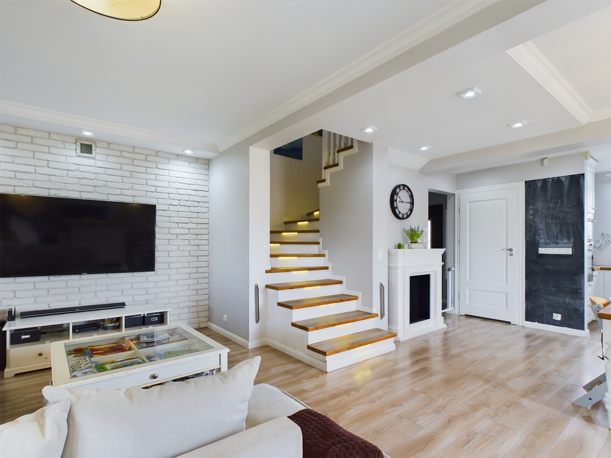 Jasne wnętrze: salon z ceglaną ścianą, telewizorem, schodami z podświetleniem, kominkiem i drewnianą podłogą. Nowoczesny design.