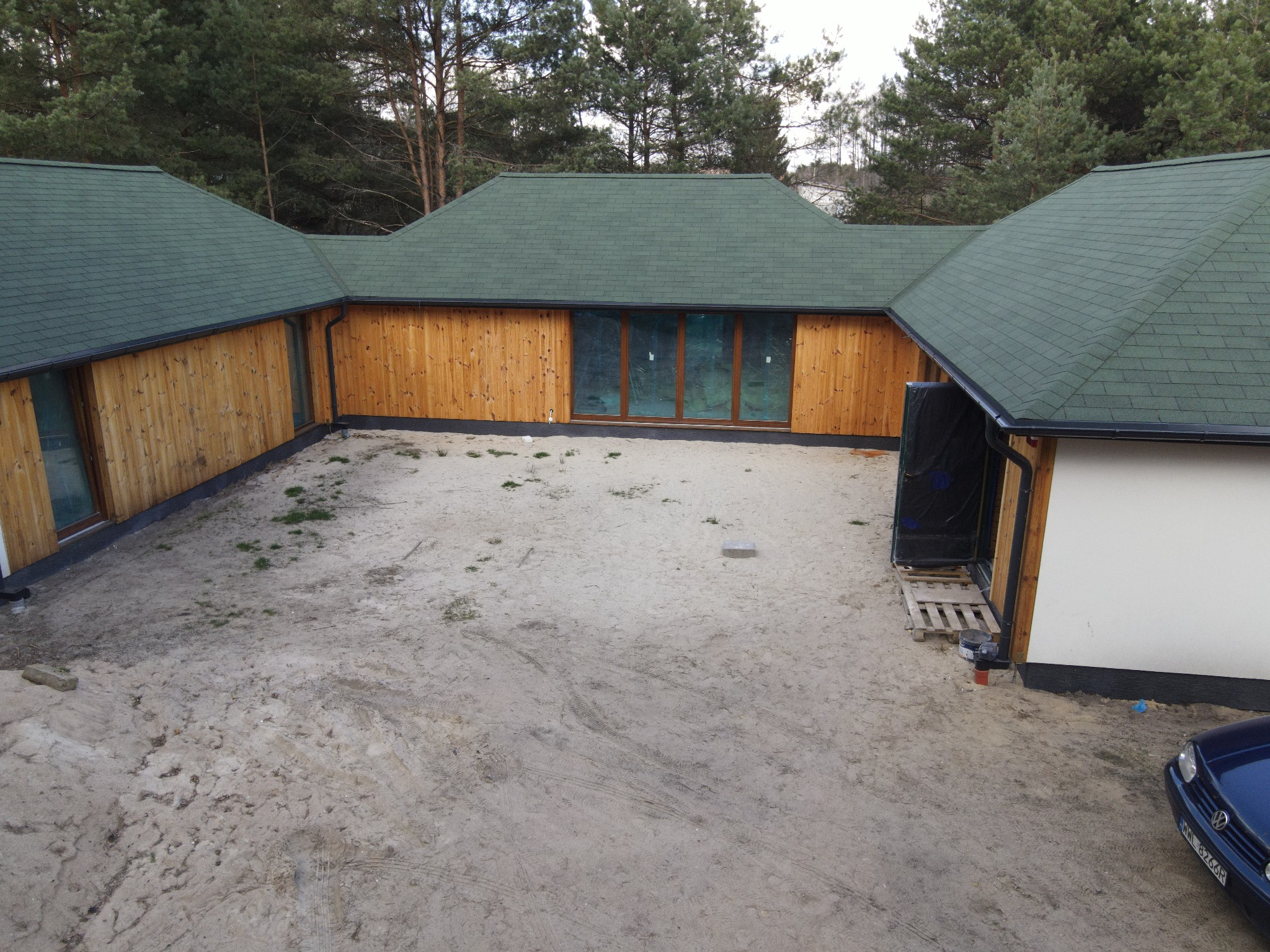 Nowoczesny, parterowy dom z drewnianą elewacją i zielonym dachem z gontu bitumicznego, otoczony piaszczystym terenem i lasem iglastym w tle.