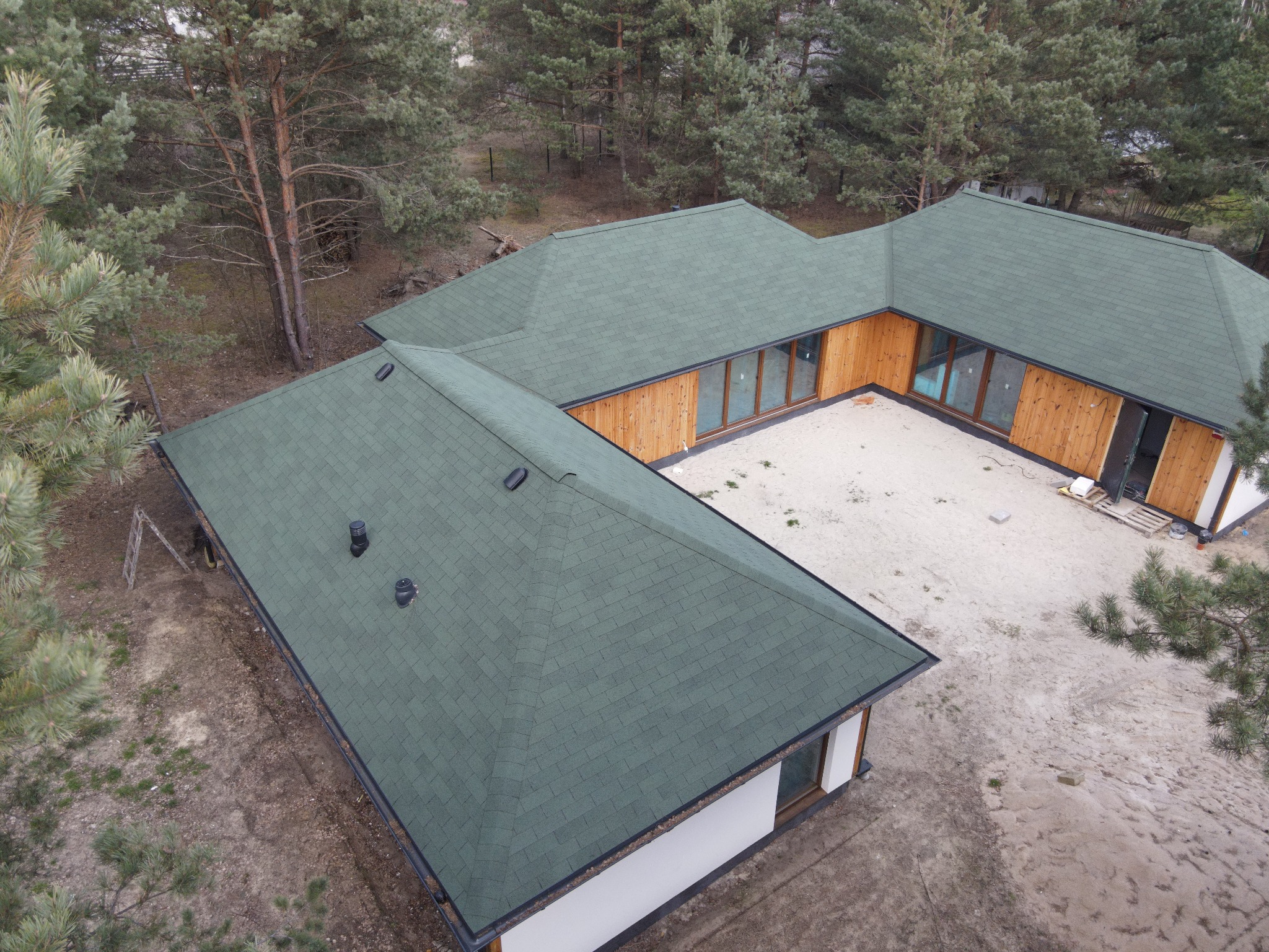 Nowoczesny dom z zielonym dachem z gontu bitumicznego, widok z góry, otoczony lasem sosnowym, w trakcie budowy, piasek na podwórku.