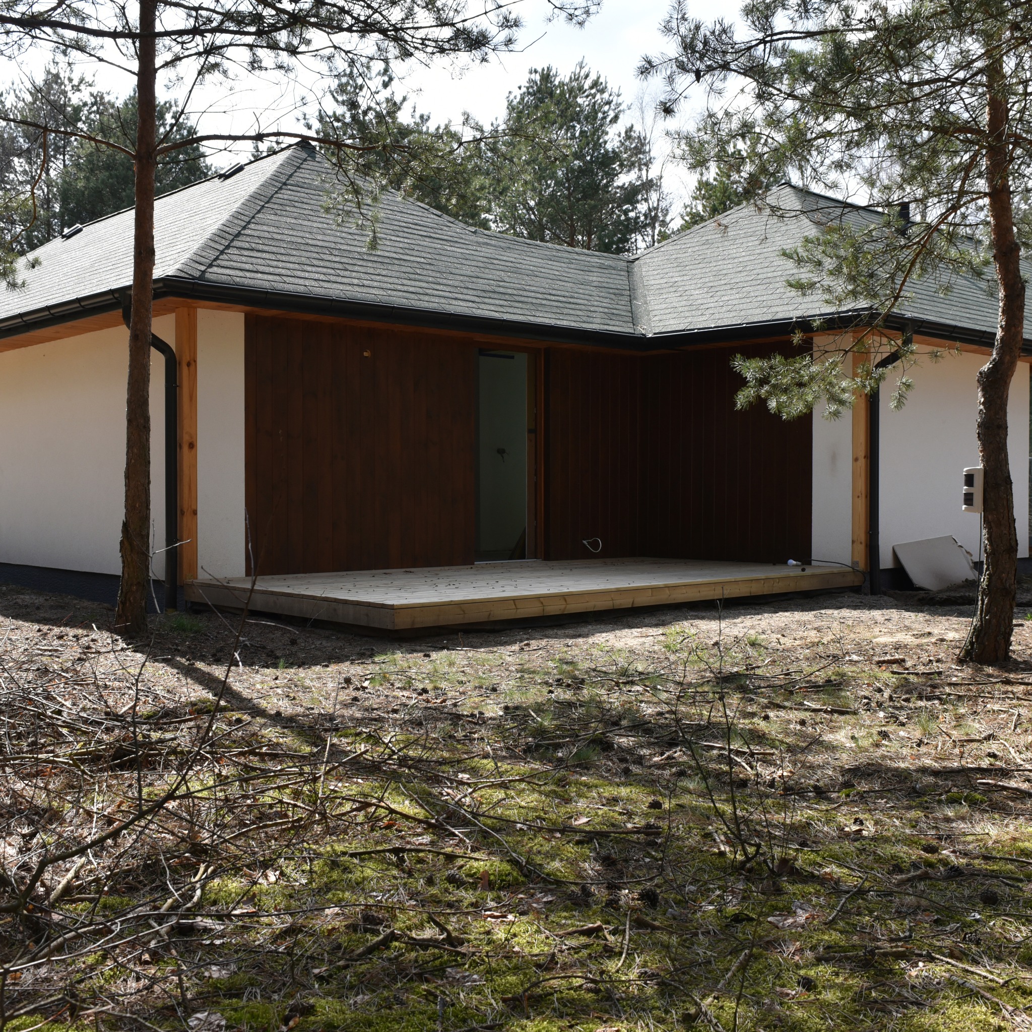 Nowoczesny dom z drewnianą elewacją i tarasem, częściowo w lesie. Widoczna surowa konstrukcja i otoczenie w naturalnym stanie.