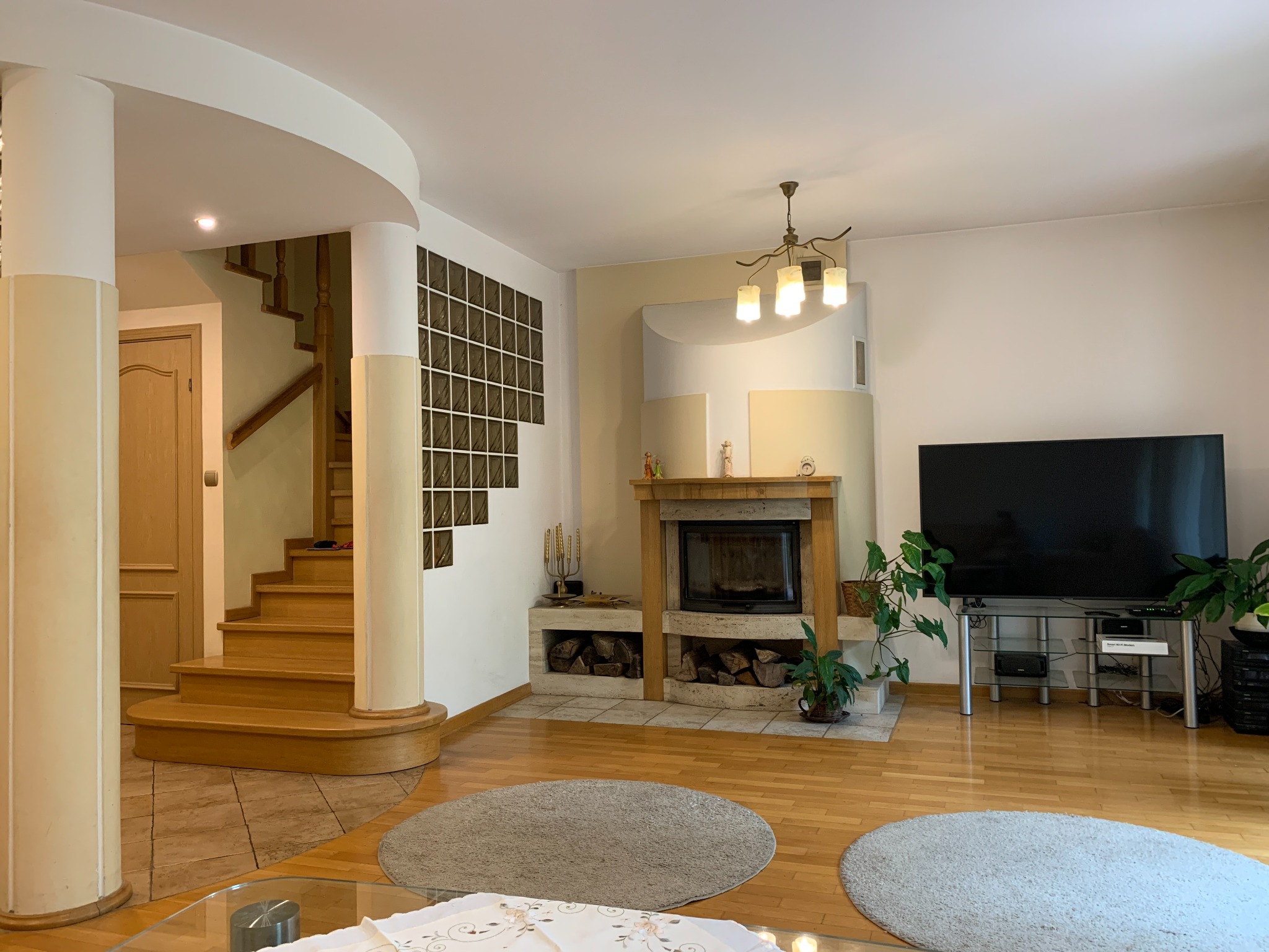Jasne wnętrze domu z drewnianymi schodami, kominkiem i telewizorem. Parkiet na podłodze, okrągłe dywany. Widok na schody i przeszkloną ścianę.