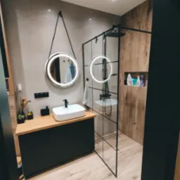 Nowoczesna łazienka z kabiną typu walk-in, okrągłym lustrem z podświetleniem LED i drewnianymi akcentami na ścianach. Minimalistyczny design.