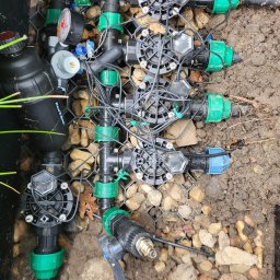 Wood Vibes Only - Złożony system nawadniania w czarnej skrzynce, z zielonymi złączkami, czarnymi rurami i przewodami, otoczony kamieniami i ziemią. Widok z góry.