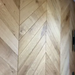 Ściana wykończona jasnym drewnem ułożonym we wzór jodełki francuskiej, z widoczną fakturą drewna i fragmentem zasłony w tle. Dekoracyjne wykończenie wnętrza.