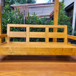 Wood Vibes Only - Drewniana ławka ogrodowa na tarasie z desek, w tle zielone krzewy i elementy architektury. Ławka o prostej konstrukcji, wykonana z jasnego drewna.