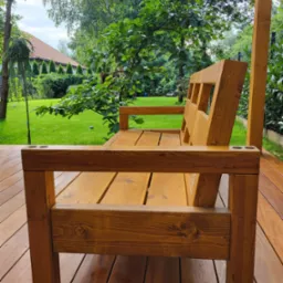 Drewniana ławka z uchwytami na napoje na tarasie z desek, w tle zielony trawnik i drzewa. Widok z boku, podkreślający solidną konstrukcję i naturalne wykończenie.