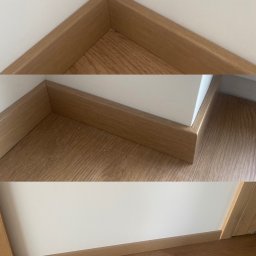 Wood Vibes Only - Zamontowane, drewnopodobne listwy przypodłogowe w rogu pomieszczenia, z widoczną strukturą drewna i drobnymi zabrudzeniami na podłodze. Białe ściany, ciepłe oświetlenie.