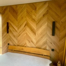 Wood Vibes Only - Drewniana ściana w jodełkę za łóżkiem, z minimalistycznymi lampami ściennymi. Widoczny fragment łóżka i stolika nocnego z dekoracyjną figurką.