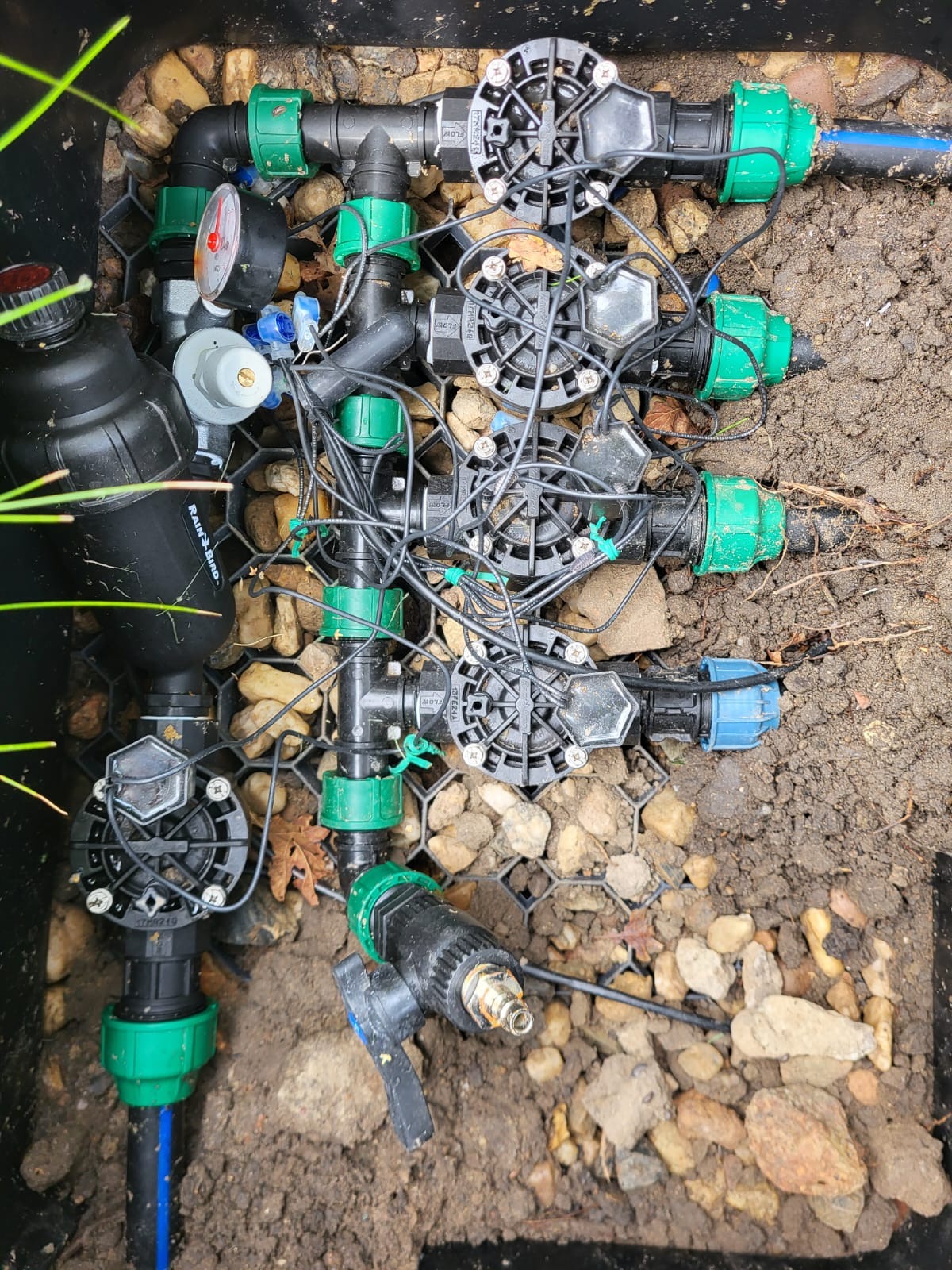 Złożony system nawadniania w czarnej skrzynce, z zielonymi złączkami, czarnymi rurami i przewodami, otoczony kamieniami i ziemią. Widok z góry.