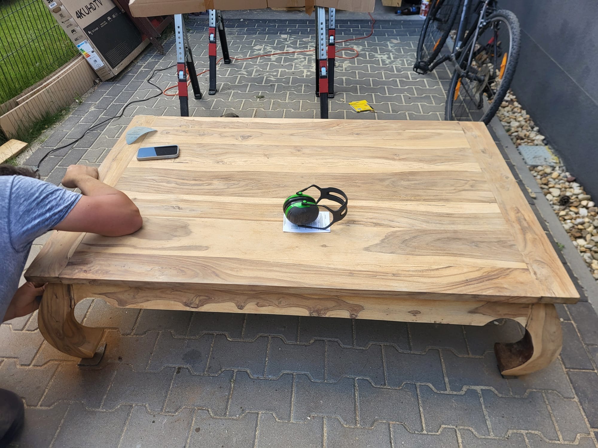 Renowacja drewnianego stolika kawowego z dekoracyjnymi nogami. Widoczne narzędzia, telefon i osoba pracująca nad wykończeniem blatu na zewnątrz.