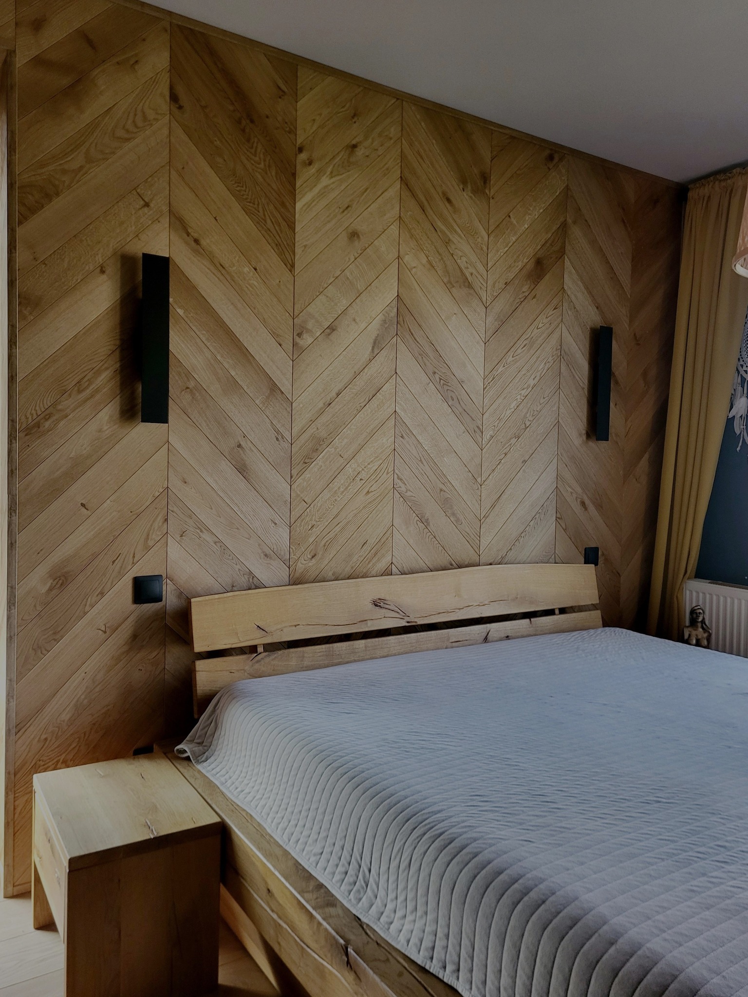 Drewniana ściana w jodełkę za łóżkiem z surowego drewna. Czarne lampy ścienne i szafka nocna z tego samego materiału. Minimalistyczny design sypialni.
