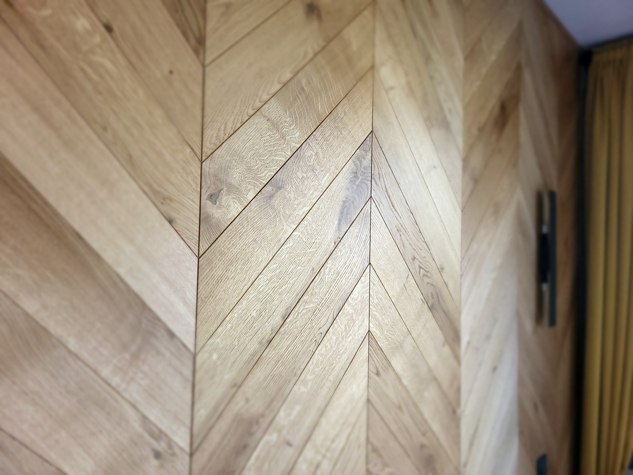 Ściana wykończona jasnym drewnem ułożonym we wzór jodełki francuskiej, z widoczną fakturą drewna i fragmentem zasłony w tle. Dekoracyjne wykończenie wnętrza.