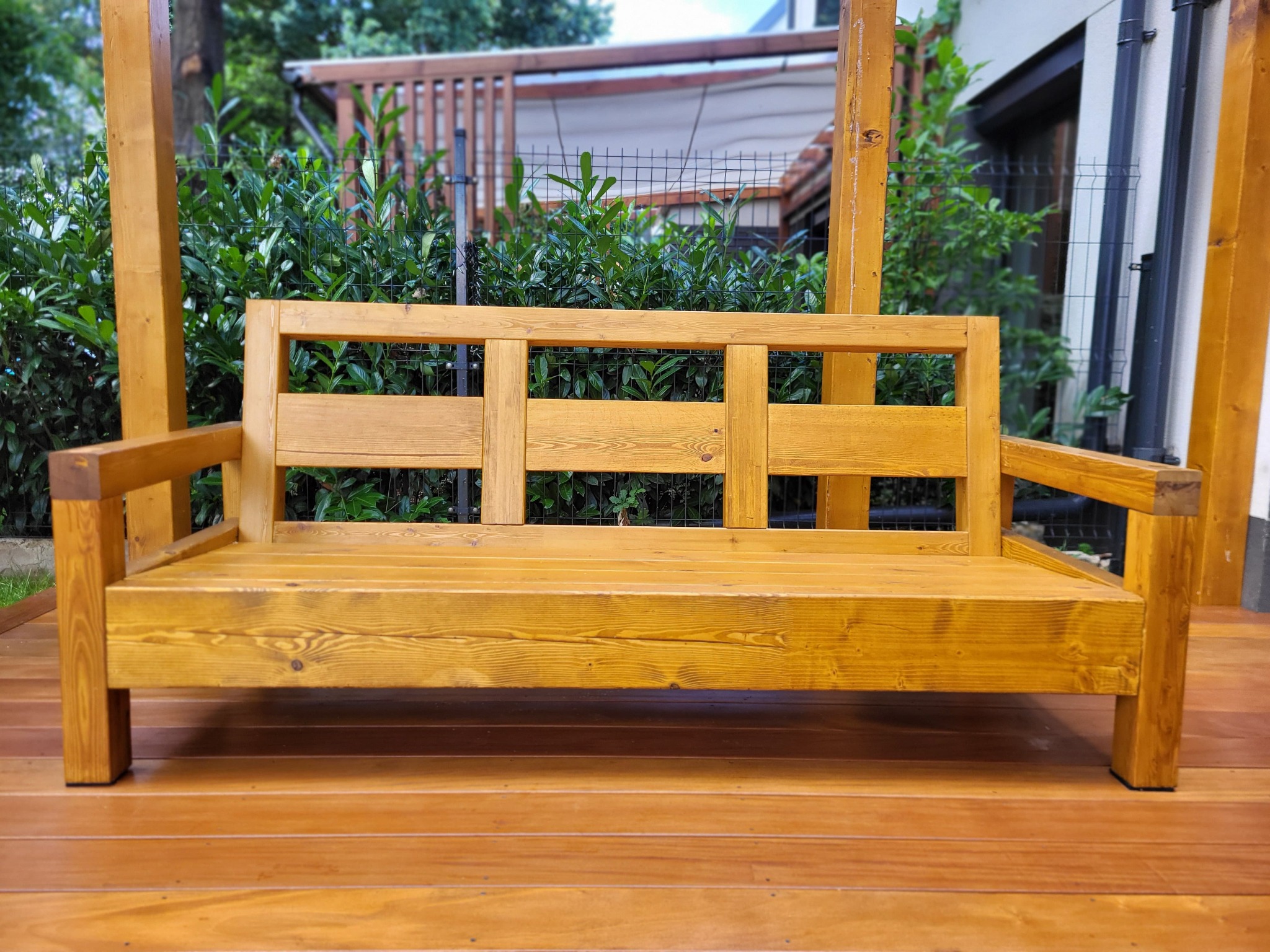 Drewniana ławka ogrodowa na tarasie z desek, w tle zielone krzewy i elementy architektury. Ławka o prostej konstrukcji, wykonana z jasnego drewna.